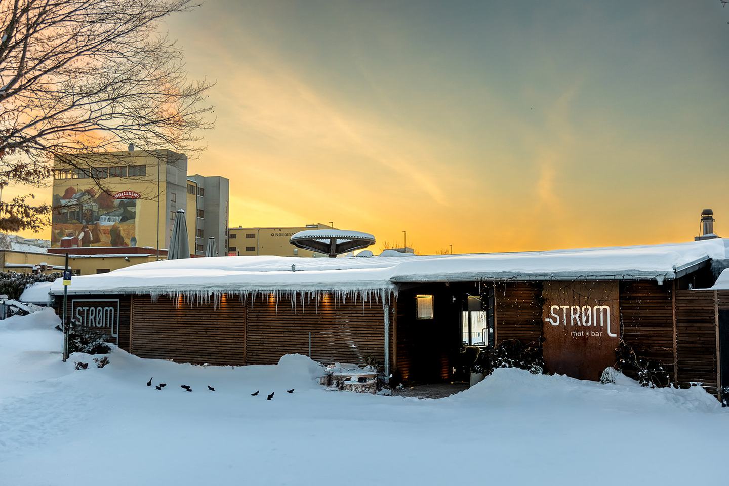 Restauranten sett utenfra en vintermorgen, med snø på taket, STRØM-skiltet på veggen og soloppgang bak
